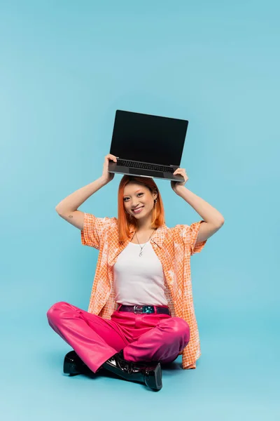 Happiness, asian woman with cheerful face and red hair holding laptop with blank screen over head while sitting with crossed legs on blue background, tattoo, young freelancer, youth culture — Stock Photo