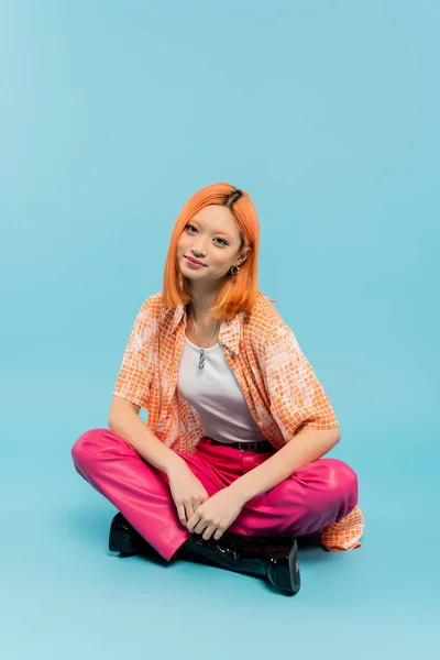 Счастливое лицо, положительные эмоции, рыжая и стильная азиатская женщина сидит со скрещенными ногами и смотрит в камеру на голубом фоне, оранжевая рубашка, розовые брюки, летние вибрации — стоковое фото