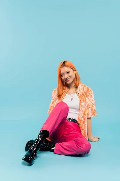 Feliz verano, joven mujer asiática con sonrisa radiante y el pelo rojo teñido mirando a la cámara en el fondo azul, atuendo casual de moda, pantalones rosas, camisa naranja, la generación z estilo de vida - foto de stock
