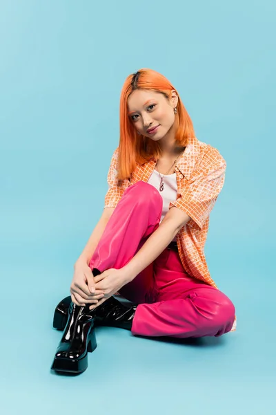 Personnalité vibrante et heureuse, pleine longueur de jeune femme asiatique à la mode assise et souriante à la caméra sur fond bleu, cheveux rouges colorés, pantalon rose, chemise orange, culture de la jeunesse — Photo de stock