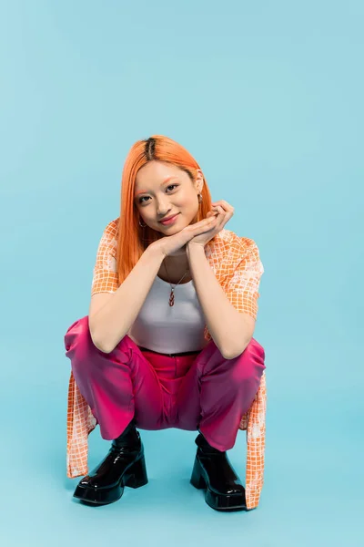 Young and stylish asian woman with pleased face expression posing on haunches and looking at camera on blue background, colored red hair, casual attire, orange shirt, pink pants, full length — Stock Photo