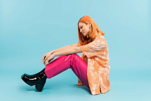 Вид сбоку молодой и стильной женщины в оранжевой рубашке и розовых брюках, сидящей на голубом фоне, окрашенные рыжие волосы, летняя мода, молодежный стиль, полный рост, поколение Z — стоковое фото