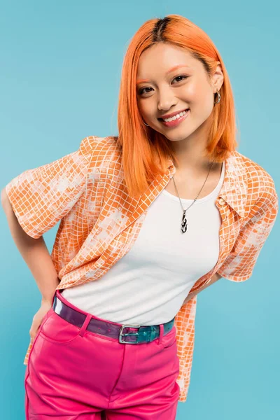 Gioiosa e alla moda donna asiatica con i capelli rossi colorati e sorriso radioso posa in camicia arancione e guardando la fotocamera su sfondo blu, felice estate, generazione z stile di vita — Foto stock
