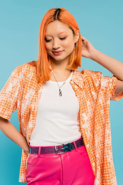 Mujer asiática joven y complacida con el pelo rojo de color rojo posando con la mano en la cadera sobre fondo azul, camisa naranja, pantalones rosados, moda juvenil, estilo veraniego, generación z - foto de stock