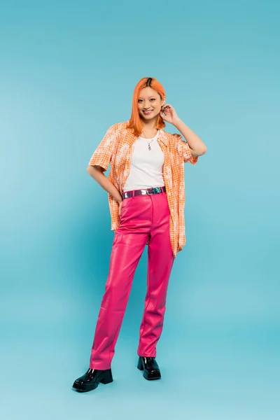 Piena lunghezza di giovane e rossa donna asiatica in abito casual alla moda in piedi con mano sul fianco e sorridente alla fotocamera su sfondo blu, emozione positiva, camicia arancione, pantaloni rosa, cultura giovanile — Foto stock
