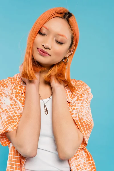 Summertime happiness, radiant smile, young and charming asian woman with colored red hair posing with closed eyes and in orange shirt on blue background, generation z — Stock Photo