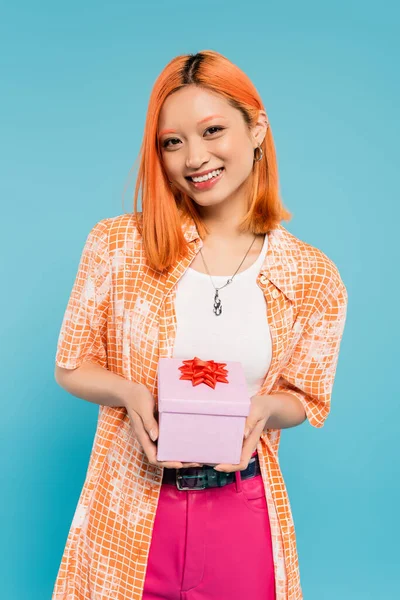 Pleasant surprise, joyful emotion, young asian woman with radiant smile holding white gift box with red ribbon on blue background, colored red hair, orange shirt, vibrant personality — Stock Photo