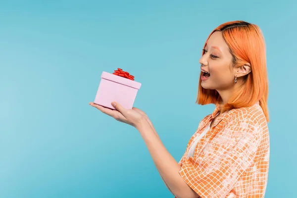 Excitement and happiness, delighted young asian woman with colored red hair and open mouth holding festive present on blue background, stylish orange shirt, youthful fashion, side view — Stock Photo