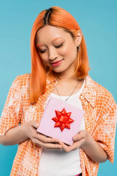 Bonheur, femme asiatique ravie en chemise orange tendance tenant boîte cadeau blanche avec arc rouge sur fond bleu, cheveux roux colorés, sourire heureux, ambiance estivale, génération z — Photo de stock