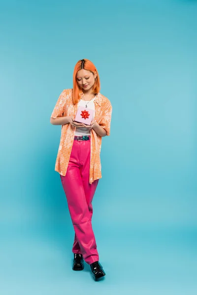 Piena lunghezza di giovane e gioiosa donna asiatica con sorriso felice, in abiti casual alla moda in piedi con scatola regalo su sfondo blu, capelli rossi tinti, camicia arancione, pantaloni rosa, moda moderna — Foto stock
