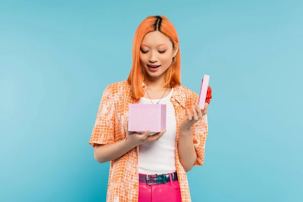 Positive emotion, young, amazed and delighted asian woman with dyed red hair, in orange shirt opening gift box with festive present on blue background, surprise, joy, happiness — Stock Photo