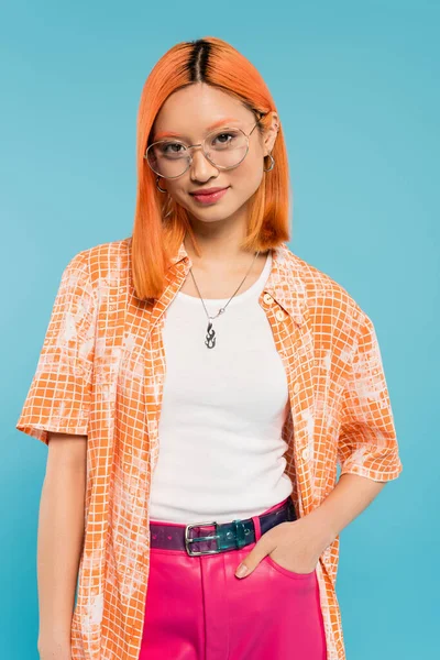 Mujer asiática feliz y de moda con el pelo rojo de color, en gafas de moda, camisa naranja y pantalones de color rosa de pie con la mano en el bolsillo mientras sonríe a la cámara en el fondo azul - foto de stock