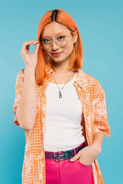 Vibrant personality, young asian woman with colored red hair adjusting stylish eyeglasses and smiling at camera on blue background, orange shirt, pink pants, fashion photography — Stock Photo