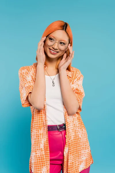 Moda de verano, felicidad, mujer asiática alegre tocando gafas de moda y mirando hacia otro lado sobre fondo azul, pelo rojo de color, camisa naranja, sonrisa radiante, estilo de vida juvenil - foto de stock