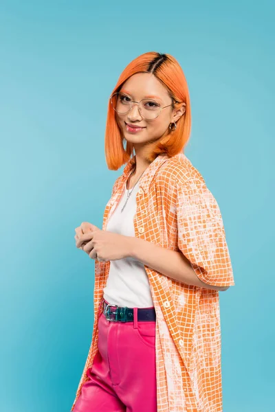Счастливое и модное лето, молодая азиатская женщина с довольным лицом, глядя в камеру, стоя на синем фоне, окрашенные рыжие волосы, стильные очки, оранжевая рубашка, поколение z образ жизни — стоковое фото