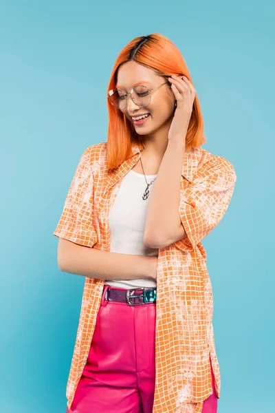 Emoción positiva, joven y alegre mujer asiática ajustando el pelo rojo de color y sonriendo sobre fondo azul, gafas de moda, camisa naranja, individualidad vibrante, moda de verano - foto de stock