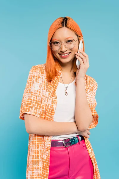 Llamada telefónica, emoción positiva, mujer asiática joven en ropa casual de moda sonriendo a la cámara durante la conversación en el teléfono inteligente sobre fondo azul, pelo rojo de color, camisa naranja, gafas con estilo - foto de stock
