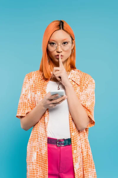 Secret, silencieux s'il vous plaît, jeune femme asiatique tenant smartphone, regardant la caméra et montrant signe de silence sur fond bleu, cheveux rouges teints, lunettes à la mode, chemise orange, mode d'été — Photo de stock
