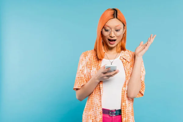 Изумление, молодая азиатская женщина с рыжими волосами и открытым ртом, глядя на мобильный телефон и жесты на синем фоне, модные очки, оранжевая рубашка, юношеская мода, цифровой образ жизни — стоковое фото