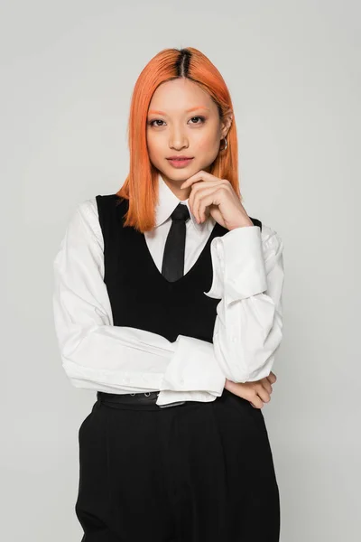 Business casual clothes, portrait of captivating asian woman with colored red hair, in white shirt, black tie and vest holding hand near face and looking at camera on grey background — Stock Photo