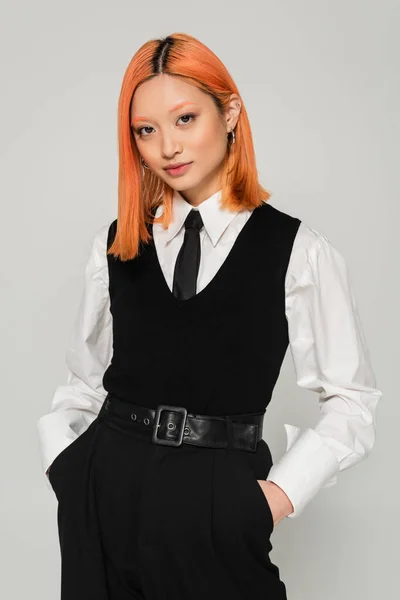 Portrait of charming and young asian woman holding hands in pockets and looking at camera on grey background, dyed red hair, white shirt, black tie, vest and pants, business fashion photography — Stock Photo