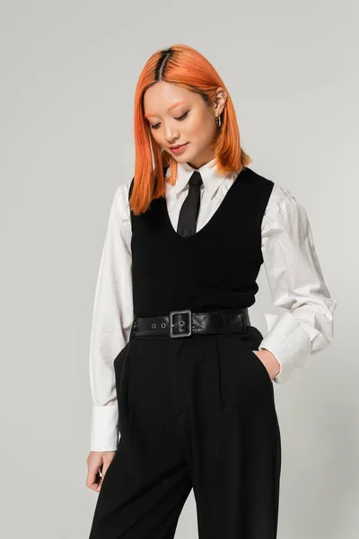 Stylish and alluring asian woman with dyed red hair, in white shirt, black tie, vest and pants posing with hand in pocket on grey background, generation z lifestyle, business casual fashion — Stock Photo