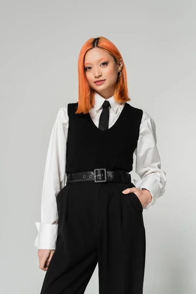 Attractive asian woman with expressive gaze holding hand in pocket and looking at camera on grey background, dyed red hair, white shirt, black tie, vest and pants, business fashion, youth culture — Stock Photo