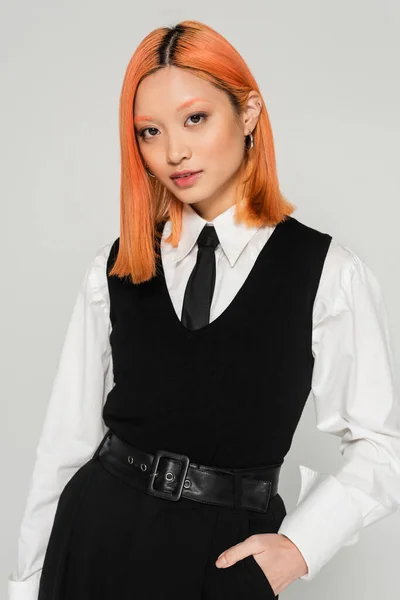 Beautiful and trendy asian woman with dyed red hair, in white shirt, black tie, vest and pants holding hand in pocket and looking at camera on grey background, business casual, youth culture — Stock Photo