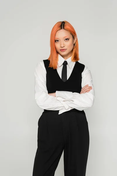 Youthful and self-assured asian woman standing with crossed arms and looking at camera on grey background, colored red hair, white shirt, black tie and pants, youthful fashion, business casual — Stock Photo