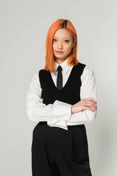 Портрет привлекательной и молодой азиатской женщины в белой рубашке, черном галстуке, жилете и брюках, стоящих со сложенными руками на сером фоне, крашеные рыжие волосы, уверенный взгляд, глядя в камеру, деловая случайность — стоковое фото