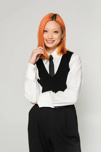 Feliz emoción, joven y alegre mujer asiática con el pelo rojo teñido mirando a la cámara sobre fondo gris, camisa blanca, corbata negra, chaleco y pantalones, fotografía de moda casual de negocios, estilo de vida moderno - foto de stock