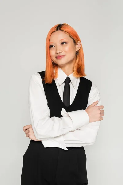Happiness, pretty and smiling asian woman standing with folded arms and looking away on grey background, colored red hair, white shirt, black vest and tie, business fashion photography — Stock Photo