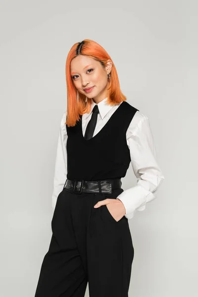 Positive and confident asian woman with colored hair holding hand in pocket and looking at camera on grey background, black and white clothes, business fashion, generation z — Stock Photo
