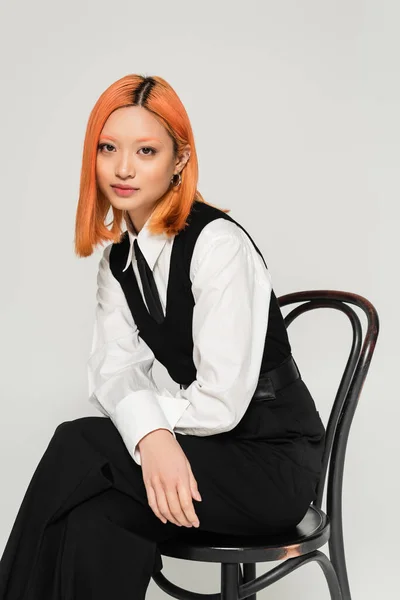 Charmante und junge asiatische Frau mit gefärbten roten Haaren sitzt auf Stuhl und blickt in die Kamera auf grauem Hintergrund, Business Casual Fashion, weißes Hemd, schwarze Krawatte und Weste, Generation Z Lifestyle — Stockfoto