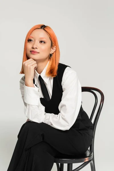 Émotion positive, visage souriant, femme asiatique rêveuse avec des cheveux rouges colorés assis sur la chaise et regardant loin sur fond gris, chemise blanche, gilet noir, vêtements décontractés d'affaires, tournage de mode — Photo de stock