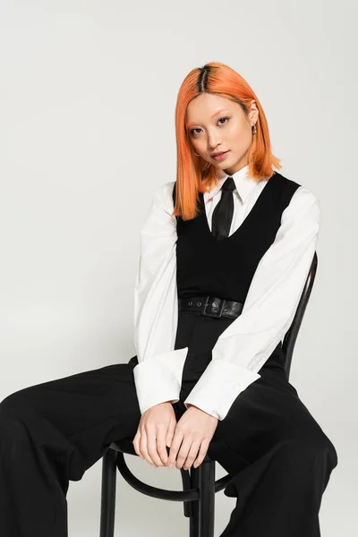 Generation z lifestyle, young asian woman with colored red hair looking at camera while sitting on chair in white shirt, black tie, vest and pants on grey background, business casual fashion shoot — Stock Photo