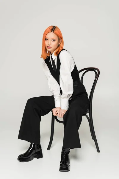 Модная фотосессия соблазнительной и трогательной женщины, смотрящей на камеру, сидя на стуле на сером фоне, окрашенные рыжие волосы, черно-белый бизнес casual, поколение Z — стоковое фото