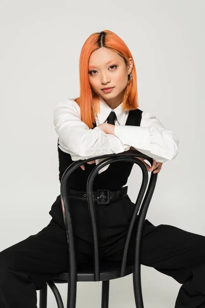Schöne und modische asiatische Frau blickt in die Kamera und posiert auf Stuhl auf grauem Hintergrund, gefärbte rote Haare, weißes Hemd, schwarze Weste und Hose, Business Casual Fashion, moderner Lebensstil — Stockfoto