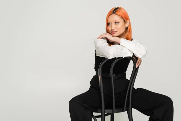 Young asian woman with colored red hair sitting on chair in black and white business casual clothes and looking away on grey background, modern fashion, generation z lifestyle — Stock Photo