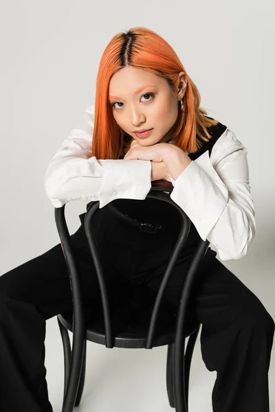 Youthful and pretty asian woman with dyed red hair wearing white shirt, black vest and pants while sitting on chair and looking at camera on grey background, business fashion photography — Stock Photo