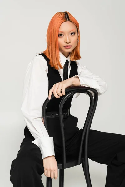 Ausdrucksstarke asiatische Frau mit gefärbten roten Haaren auf Stuhl sitzend und vor der Kamera posierend auf grauem Hintergrund, gefärbte rote Haare, schwarz-weiße Kleidung, Business-Modefotografie, Generation z — Stockfoto