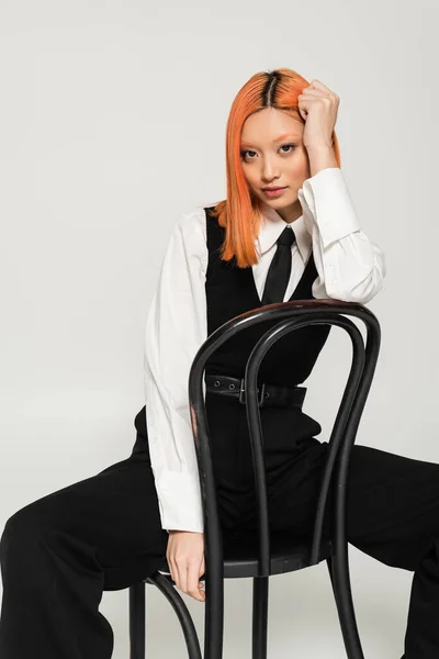 Verführerische und stilvolle asiatische Frau mit gefärbten roten Haaren, in weißem Hemd, schwarzer Krawatte, Weste und Hose, die in die Kamera schaut, während sie auf einem Stuhl auf grauem Hintergrund sitzt, Generation Z Lifestyle, Business-Mode — Stockfoto
