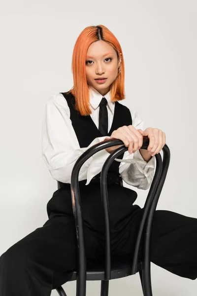 Молодая, привлекательная и рыжая азиатская женщина в белой рубашке, черном галстуке, жилете и брюках, смотрящих в камеру, сидя на стуле на сером фоне, деловом стиле, поколение z — стоковое фото
