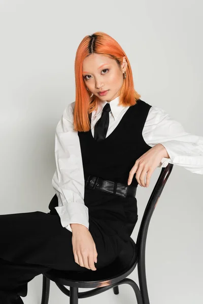 Jeune et enchanteur modèle asiatique avec des cheveux rouges regardant la caméra et assis sur la chaise sur fond gris, chemise blanche, gilet noir, cravate et pantalon, mode décontractée d'affaires, style moderne — Photo de stock