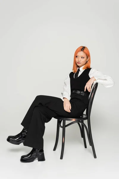 Full length of redhead asian woman in black and white casual clothes posing on chair and looking away on grey background, colored red hair, shirt, tie, vest and pants, business casual style — Stock Photo