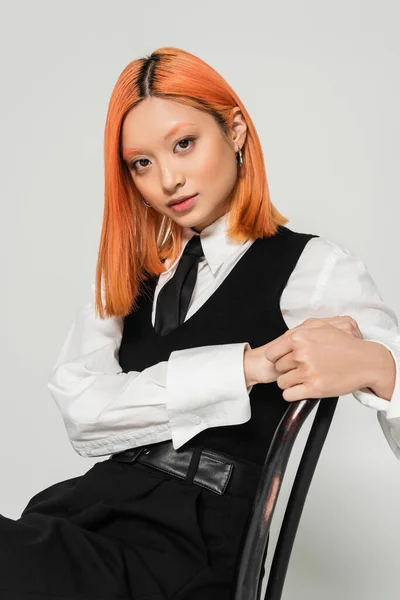 Atraente e jovem mulher asiática com cabelos vermelhos tingidos, vestindo camisa branca, colete preto e gravata, sentado na cadeira e olhando para a câmera no fundo cinza, tiro de moda, negócios casuais — Fotografia de Stock