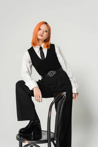 Pretty and confident asian woman in white shirt, black tie, vest and pants, with colored red hair stepping on chair and looking at camera on grey background, modern business fashion, generation z — Stock Photo