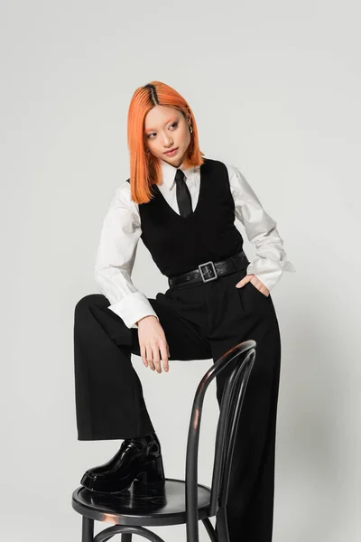Junge und selbstbewusste asiatische Frau hält die Hand in der Tasche und schaut weg, während sie mit Stuhl auf grauem Hintergrund posiert, gefärbte rote Haare, schwarz-weiße Business-Freizeitkleidung, Mode-Shooting — Stockfoto