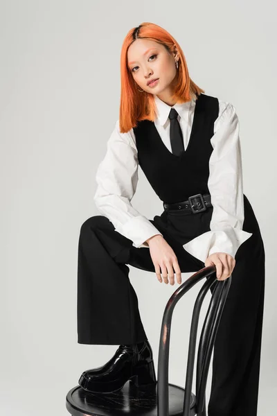 Charmante und trendige asiatische Frau, die in die Kamera schaut, wenn sie auf einem Stuhl auf grauem Hintergrund posiert, gefärbte rote Haare, schwarz-weiße Business-Freizeitkleidung, Lebensstil der Generation Z — Stockfoto