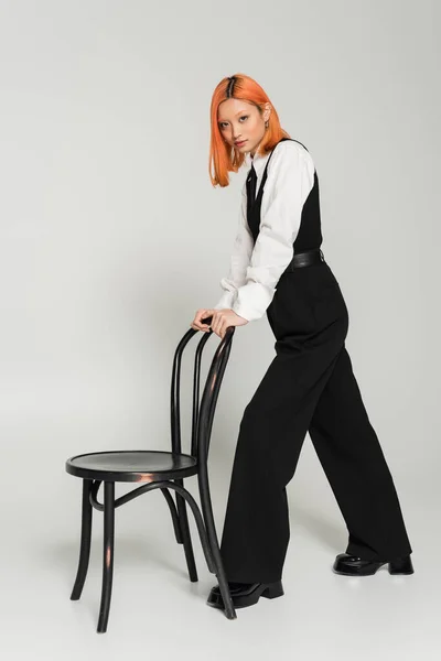 Full length of fashionable and red haired asian woman standing near chair and looking at camera on grey background, black and white clothes, business casual fashion, generation z — Stock Photo